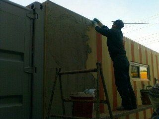Person applying stucco to a wall while standing on a ladder next to a shipping container.
