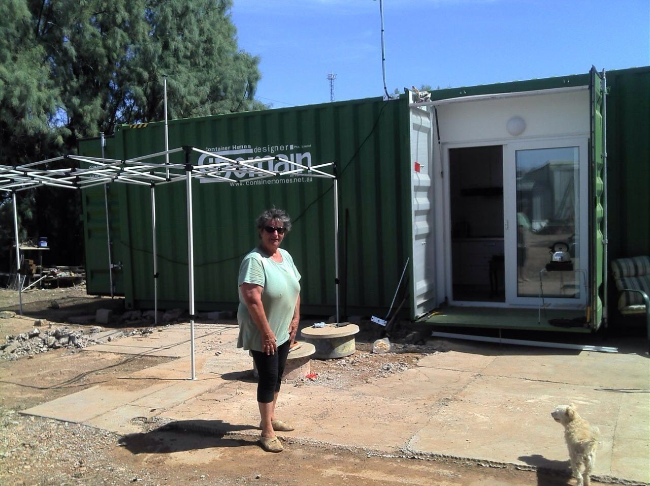 Woman standing outside a green shipping container home; small dog on the right.