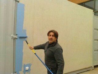 Man painting a metal container door blue with a roller. Beige container wall in background.