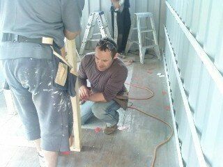 Two people inside a structure building, one kneeling with tool. Grey and blue clothing.