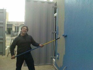Man painting blue paint on a building's exterior with a roller. Outdoors, near a metal structure.