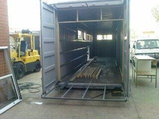 Open gray shipping container interior with partial wooden framework and windows. A forklift and van are visible nearby.