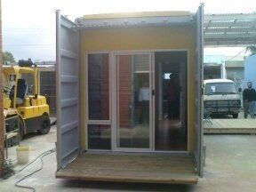 Shipping container converted into a small building with door and windows, wooden deck, and open side doors, yellow and grey.