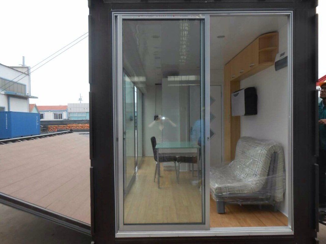 Shipping container converted into a small living space with glass sliding door; interior features furniture, wooden floors, and a dining table.
