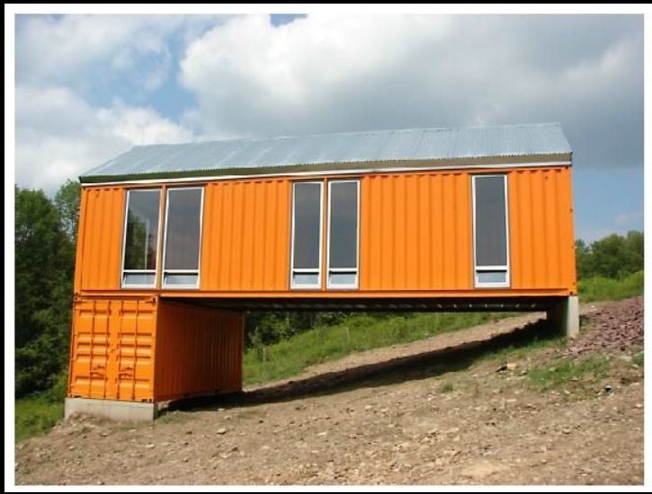 Orange shipping container home elevated over sloped terrain. Rectangular windows, metal roof, and concrete supports.