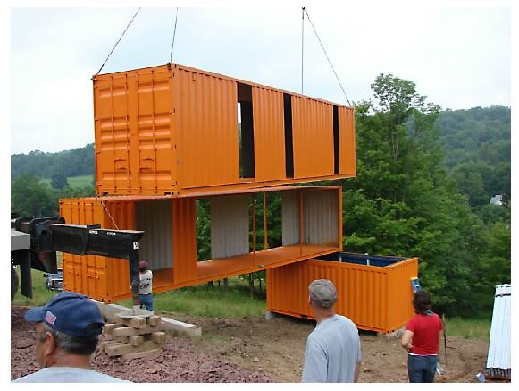Orange shipping containers being lifted by a crane to assemble a house on a grassy hillside.