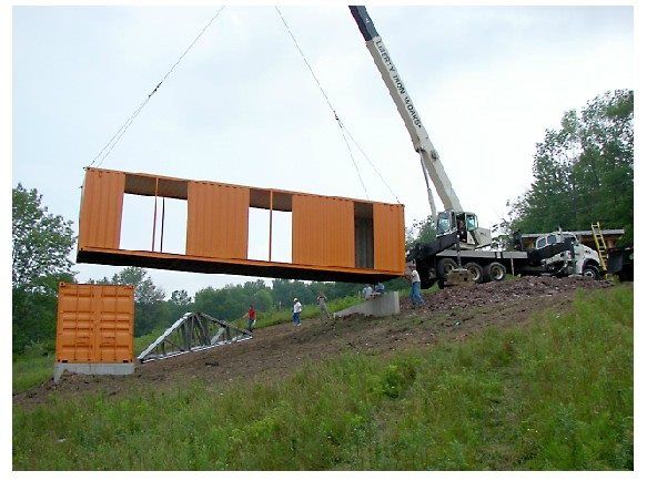Crane lifting orange shipping container with cutouts for windows and doors. Construction site outdoors.