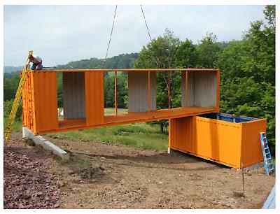 Orange shipping containers being assembled on a hillside. A worker on a ladder.