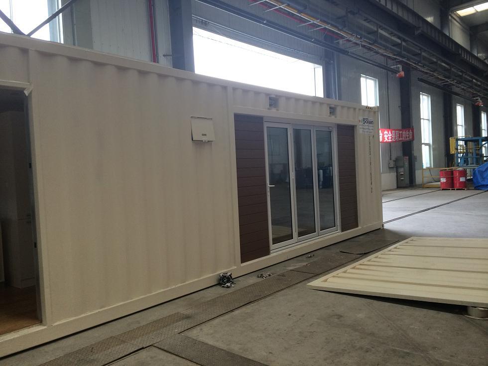 A white modular building with sliding glass doors and brown wooden panels, inside a large industrial warehouse.