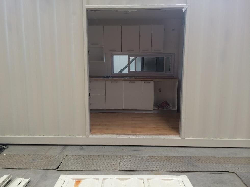 Interior view of a modular structure with a kitchen visible through an opening. White walls, cabinets, and a wooden floor.