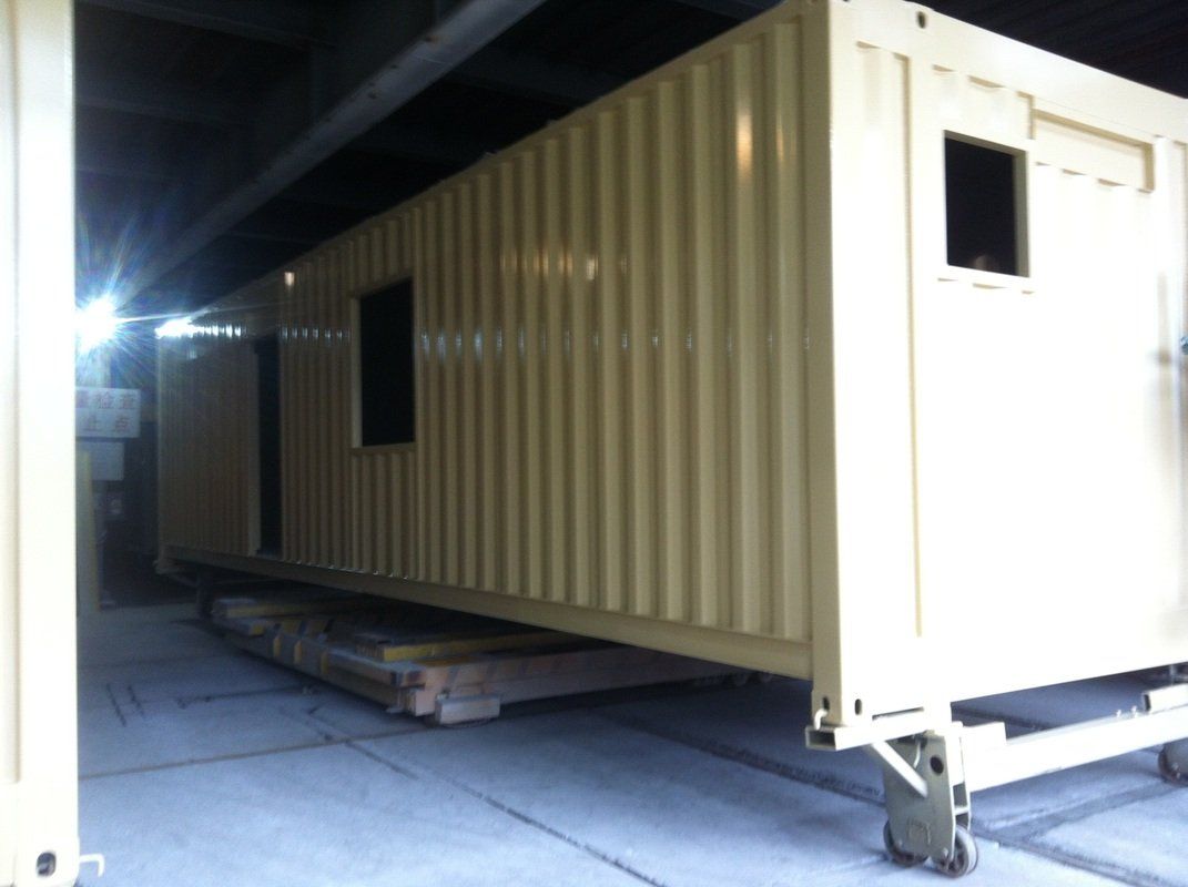 Tan rectangular shipping containers with windows and doors, on wheels under a structure.