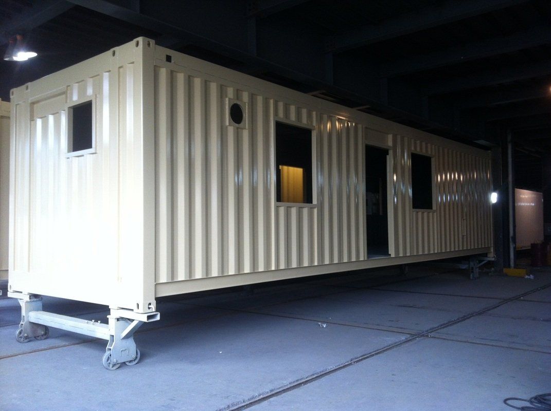Tan modified shipping container with windows and doors, on wheels, in an industrial setting.