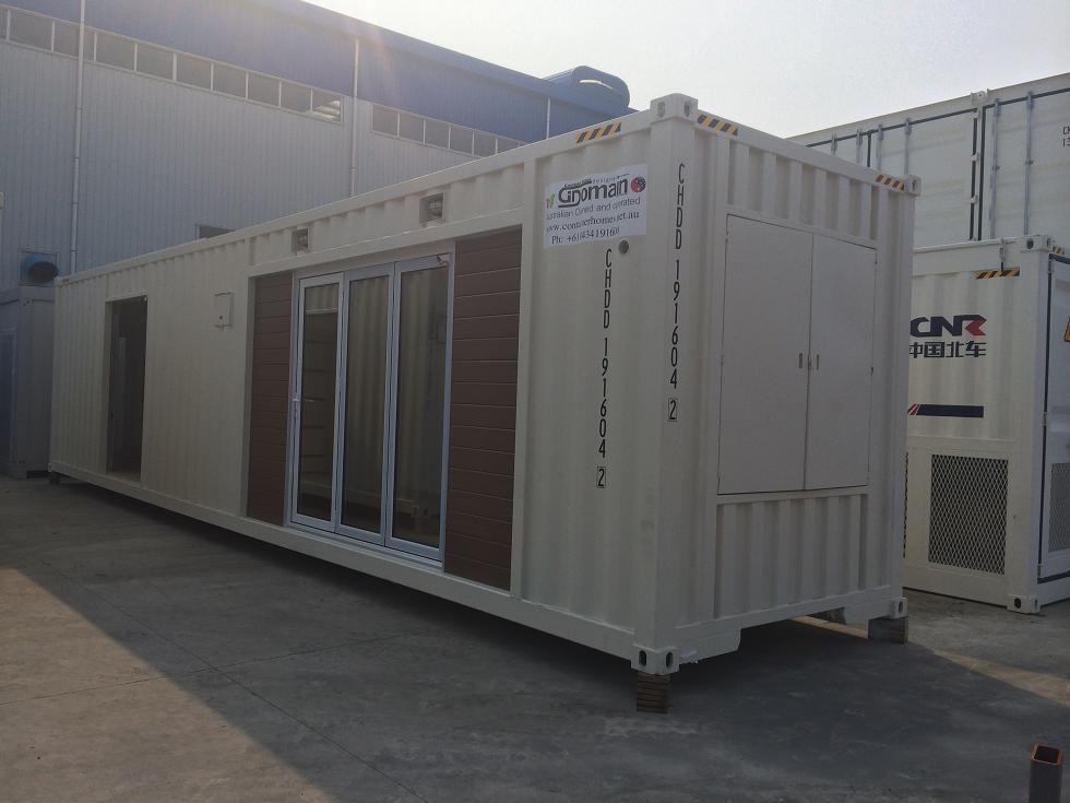 Tan shipping container modified for housing with sliding glass doors, a door, and windows, outdoors.