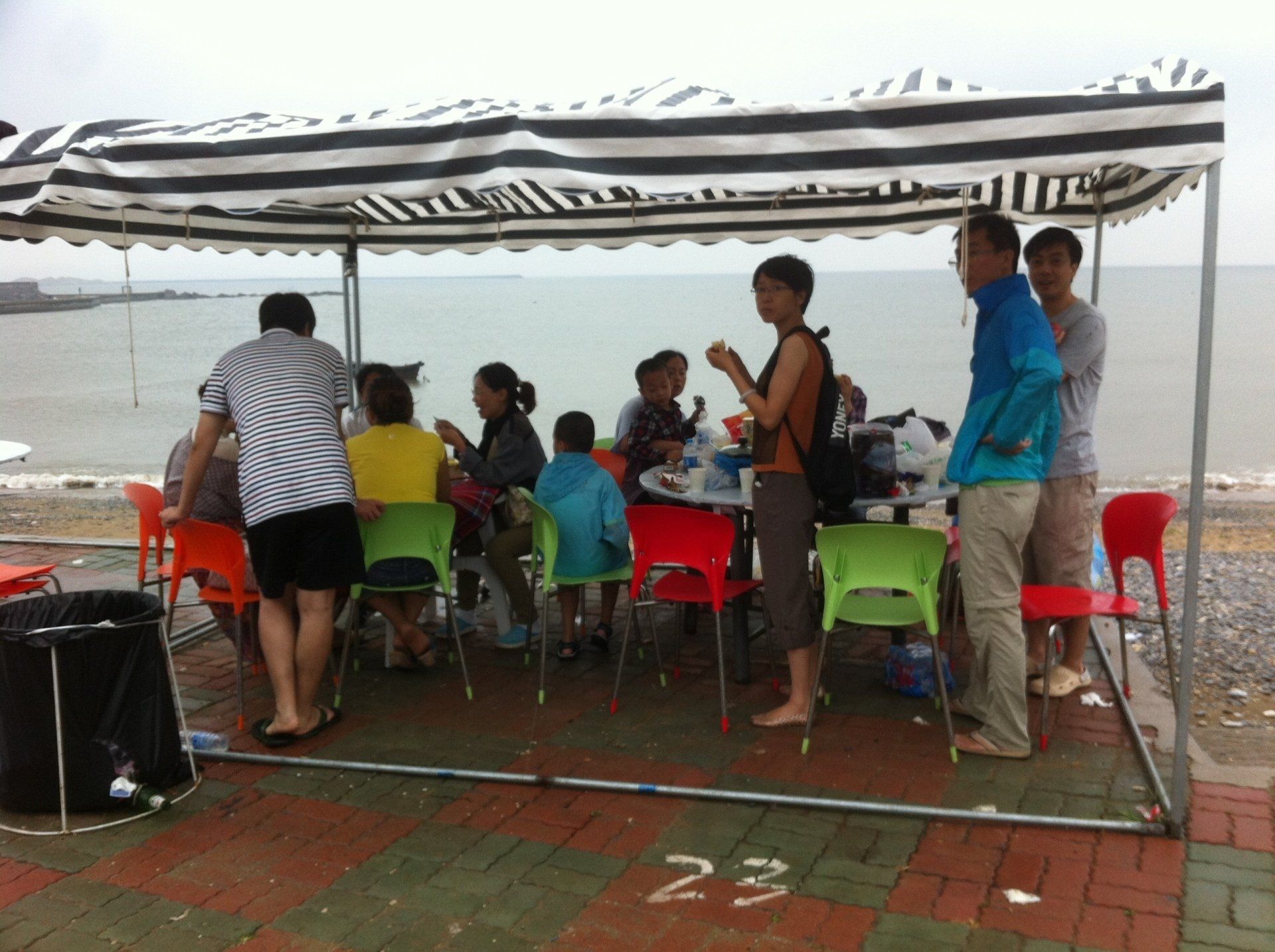 People eating at a table under a striped canopy on a beach. Red and green chairs. Ocean in background.