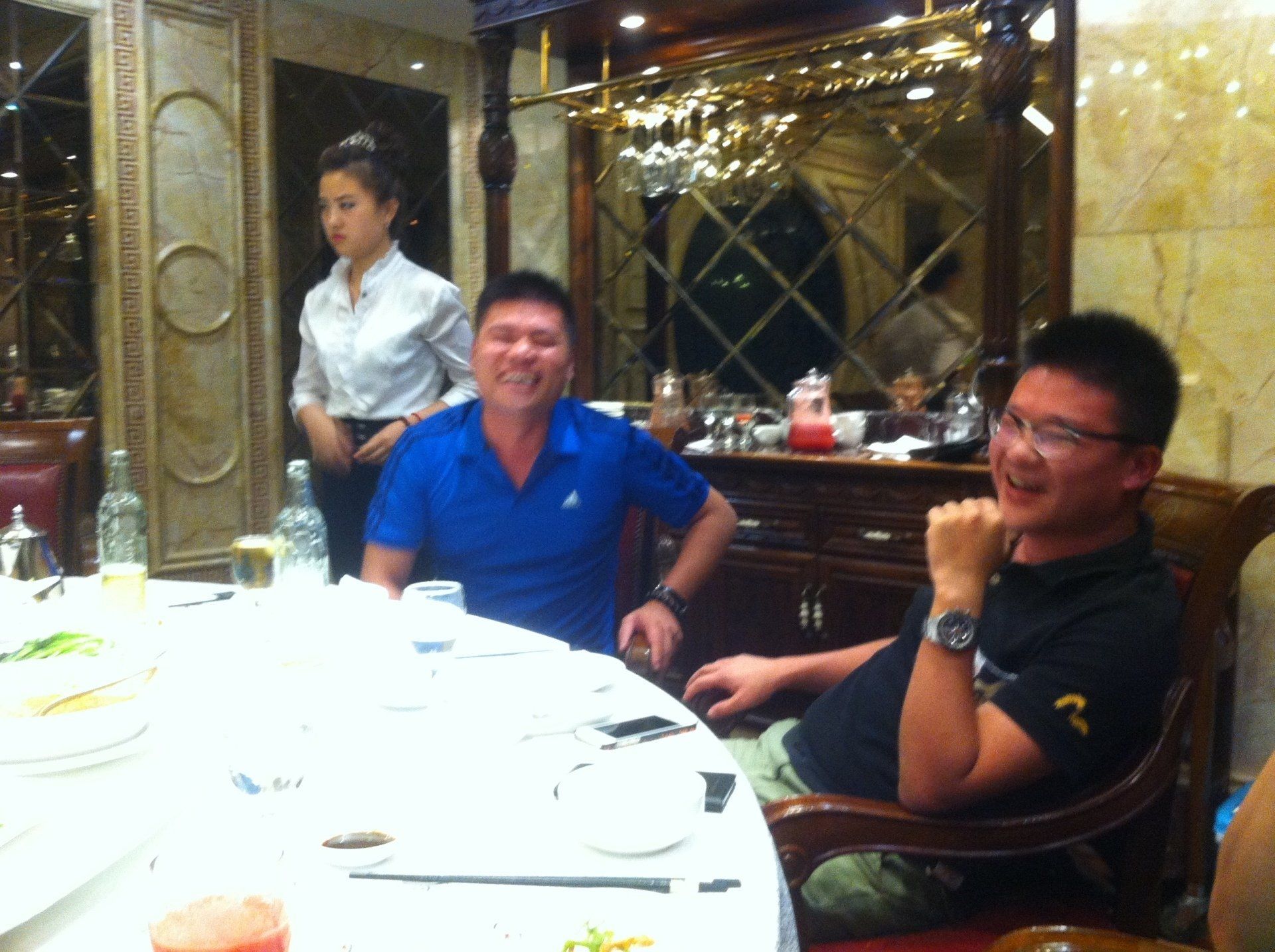 Two men seated at a round table in a restaurant; a server stands nearby. One man smiles, the other laughs.