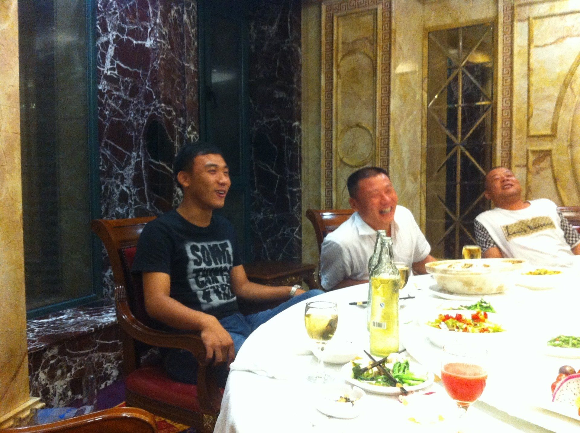 Three men laughing around a dining table set with food and drinks; ornate interior.
