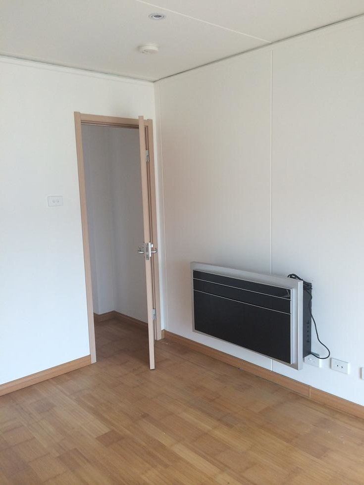 Empty room with wooden floor, white walls, open doorway, and wall-mounted black heater.