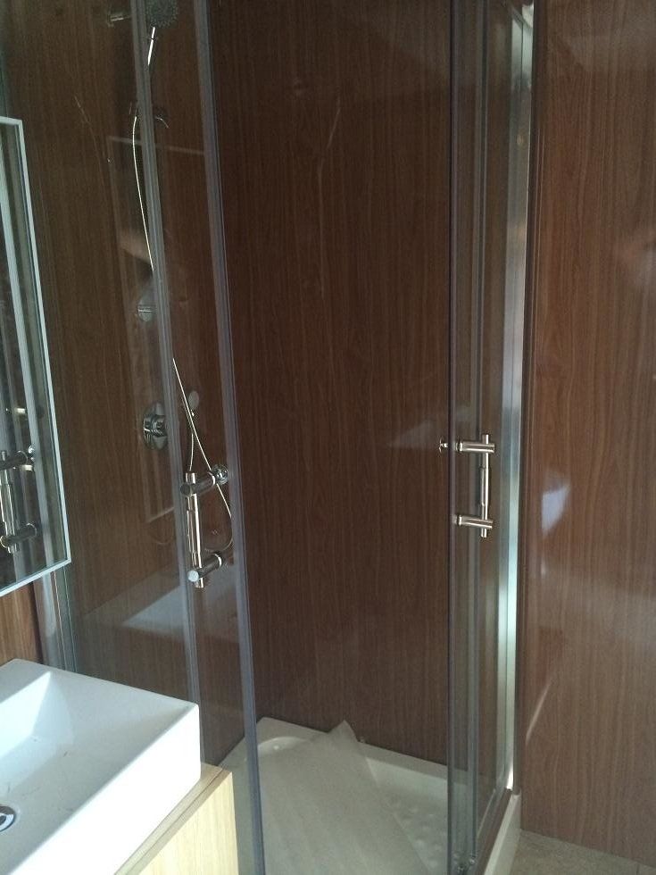 Shower stall with glass doors, silver fixtures, and brown wood-paneled walls, next to a white sink.