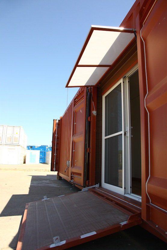 Rust-colored shipping container with open sliding glass door, ramp, and awning under blue sky.