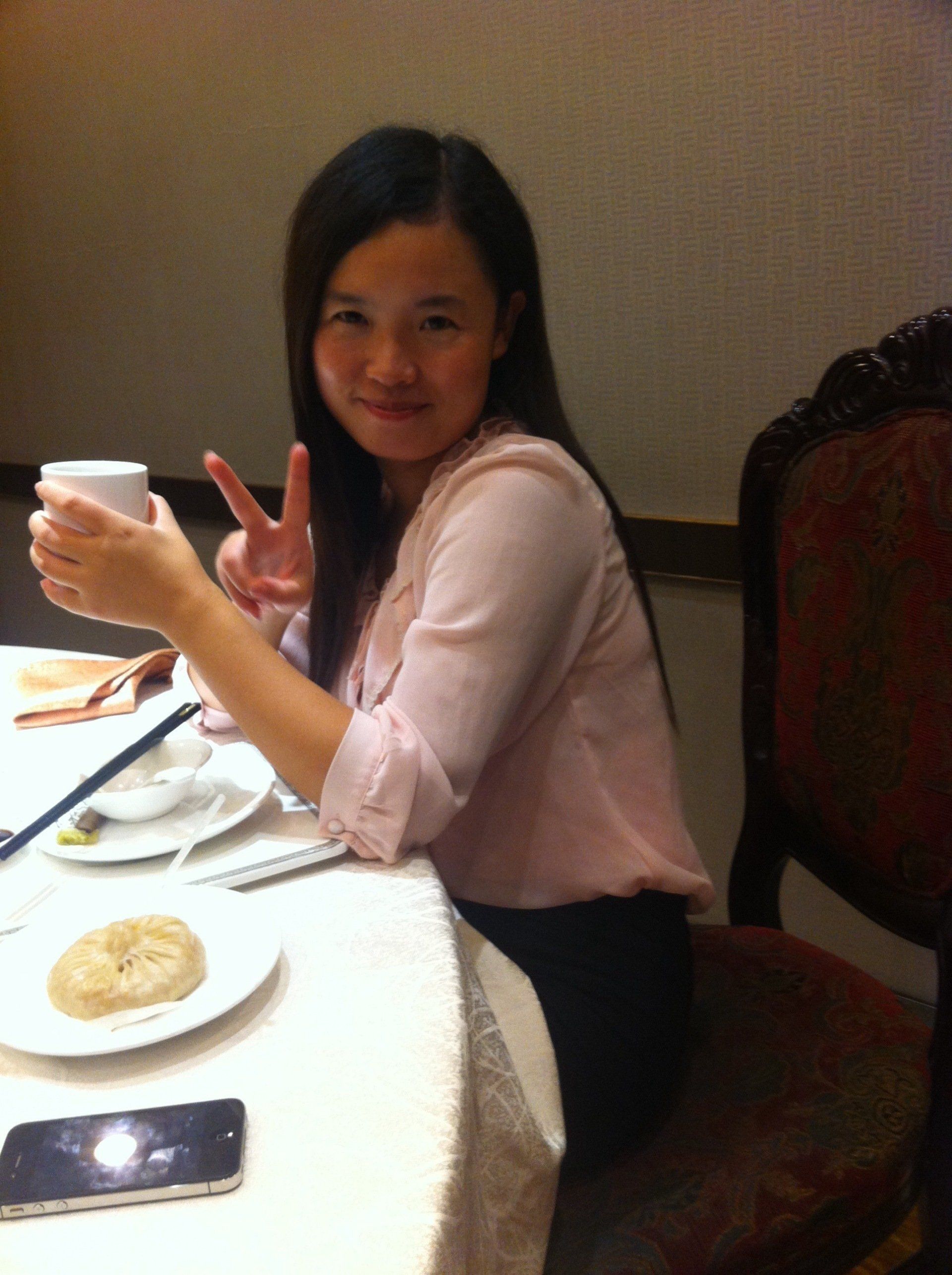 Woman at a table holding a cup, making a peace sign, smiling. Setting appears to be a restaurant.