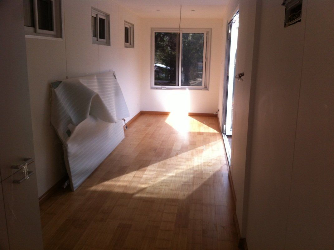 Empty room with wood floor, window with sunlight, and white walls. Packaging leans against the wall.