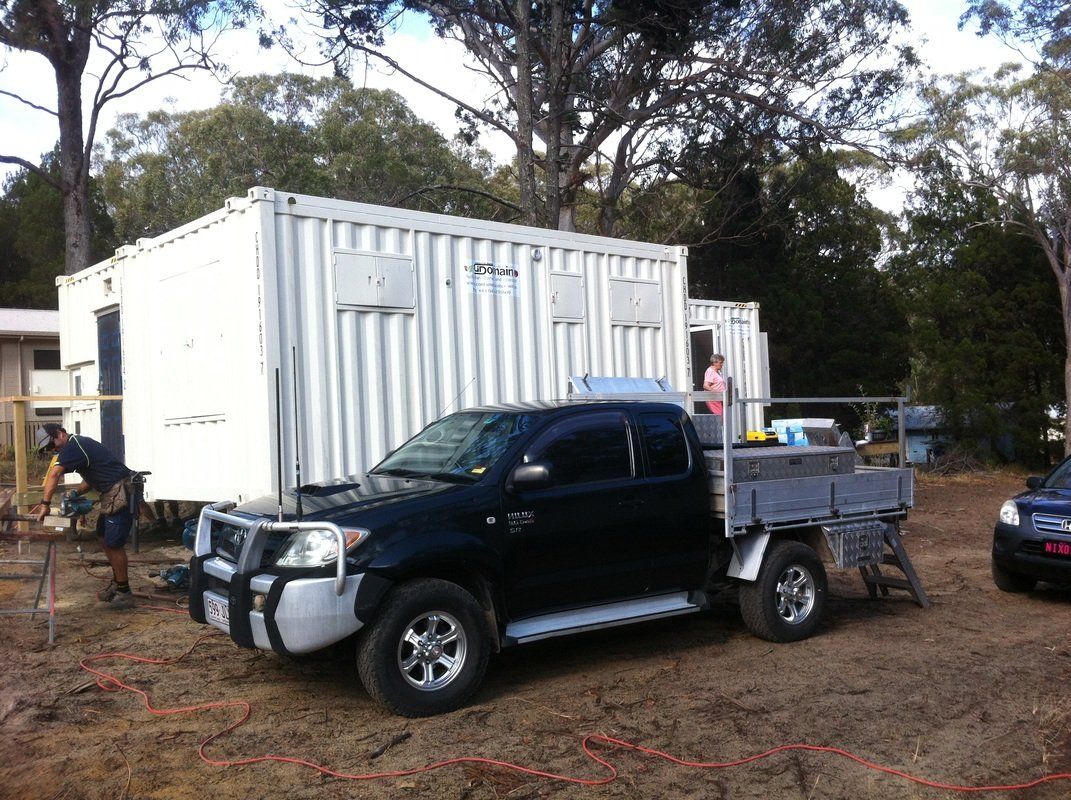 Black pickup truck parked in front of white containers, possibly used for construction, with person in background.