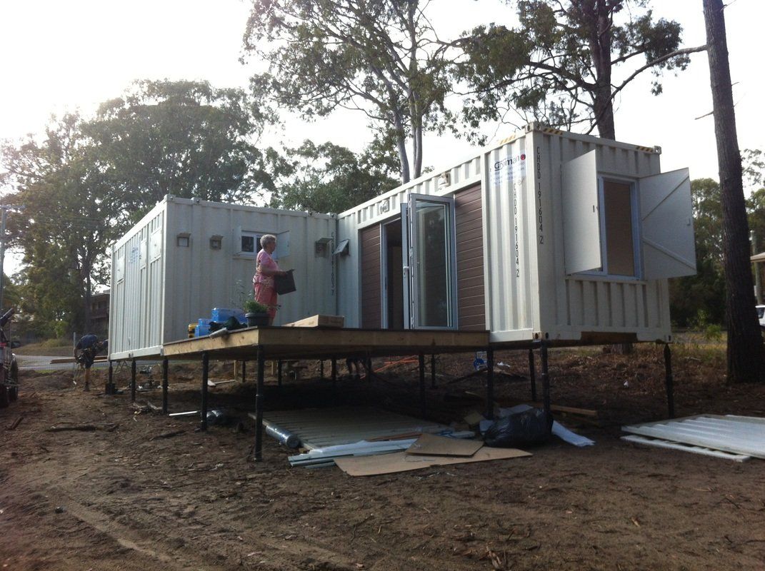 Shipping container home under construction; person on deck, trees in background.