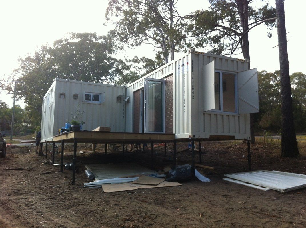 Container home under construction on stilts, doors and windows open, in a wooded area.