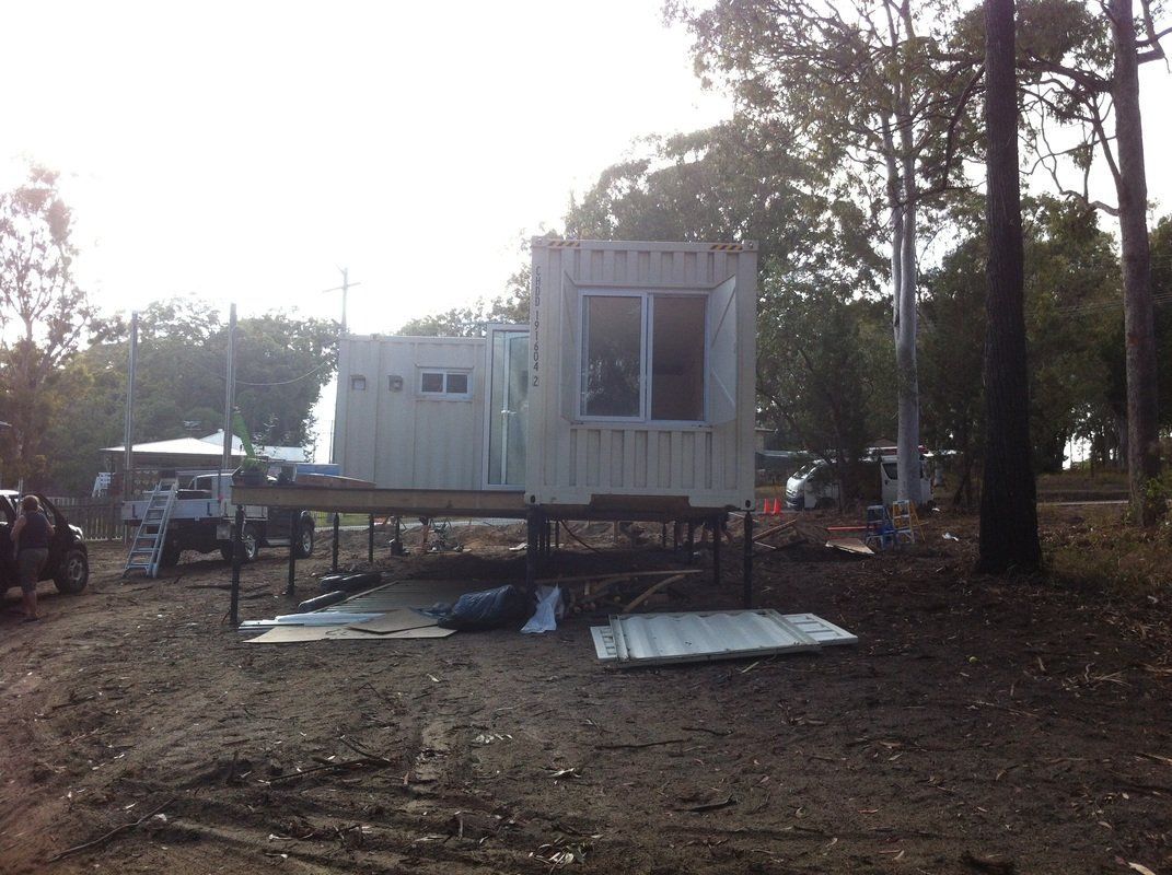 Shipping container structure on stilts in a dirt area, with trees and a truck visible in the background.