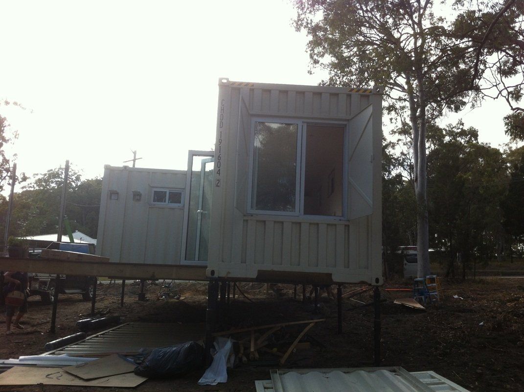 Shipping container home under construction, white walls, large window, raised platform.
