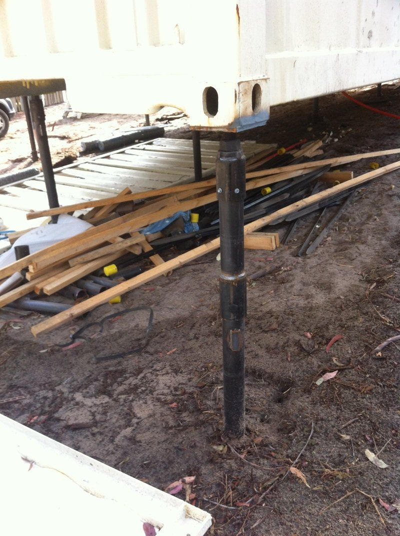 Shipping container elevated on adjustable supports over dirt and debris.