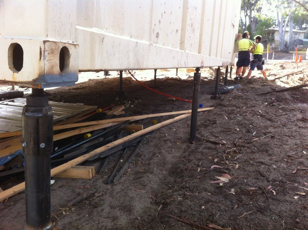 Construction site. Large white structure raised on jacks. Two workers in vests, dirt ground.