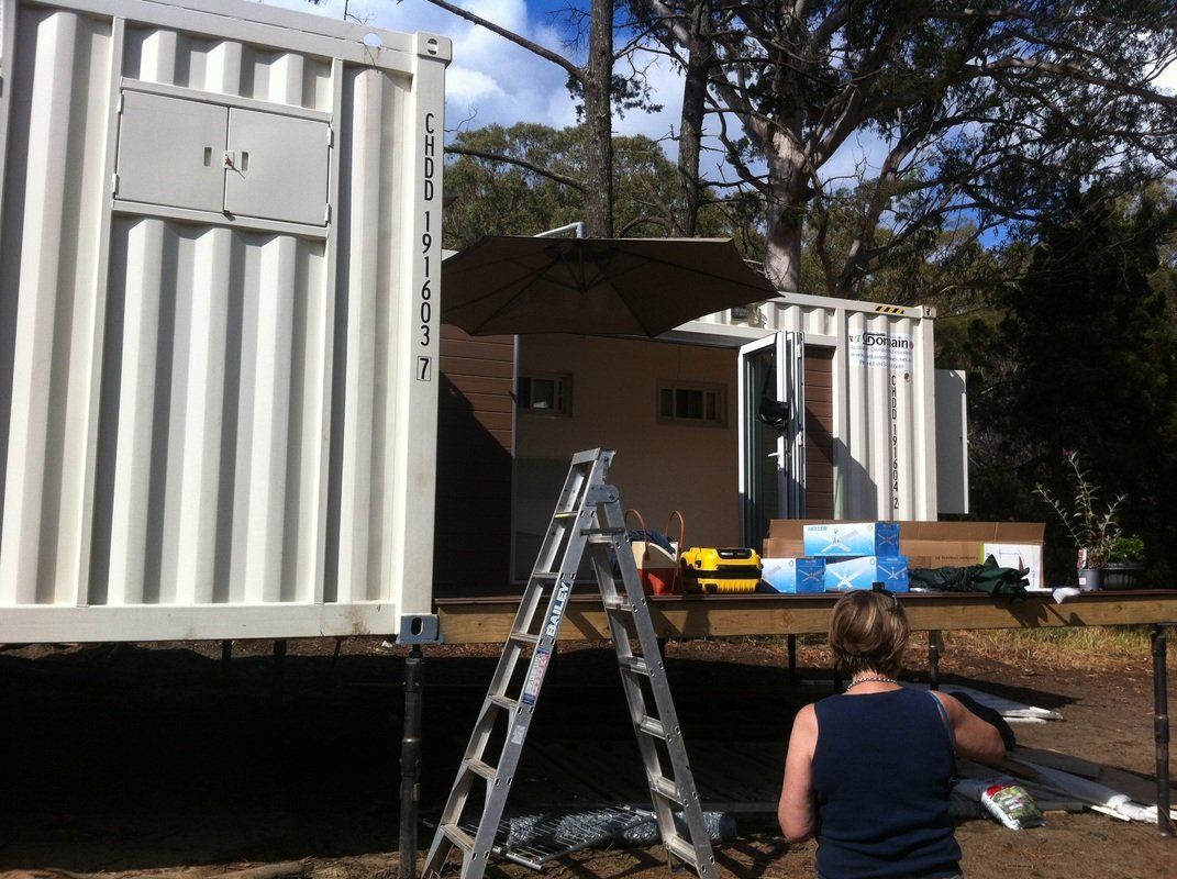 Construction site with shipping containers, a woman working, ladder, and umbrella in sunny outdoor setting.