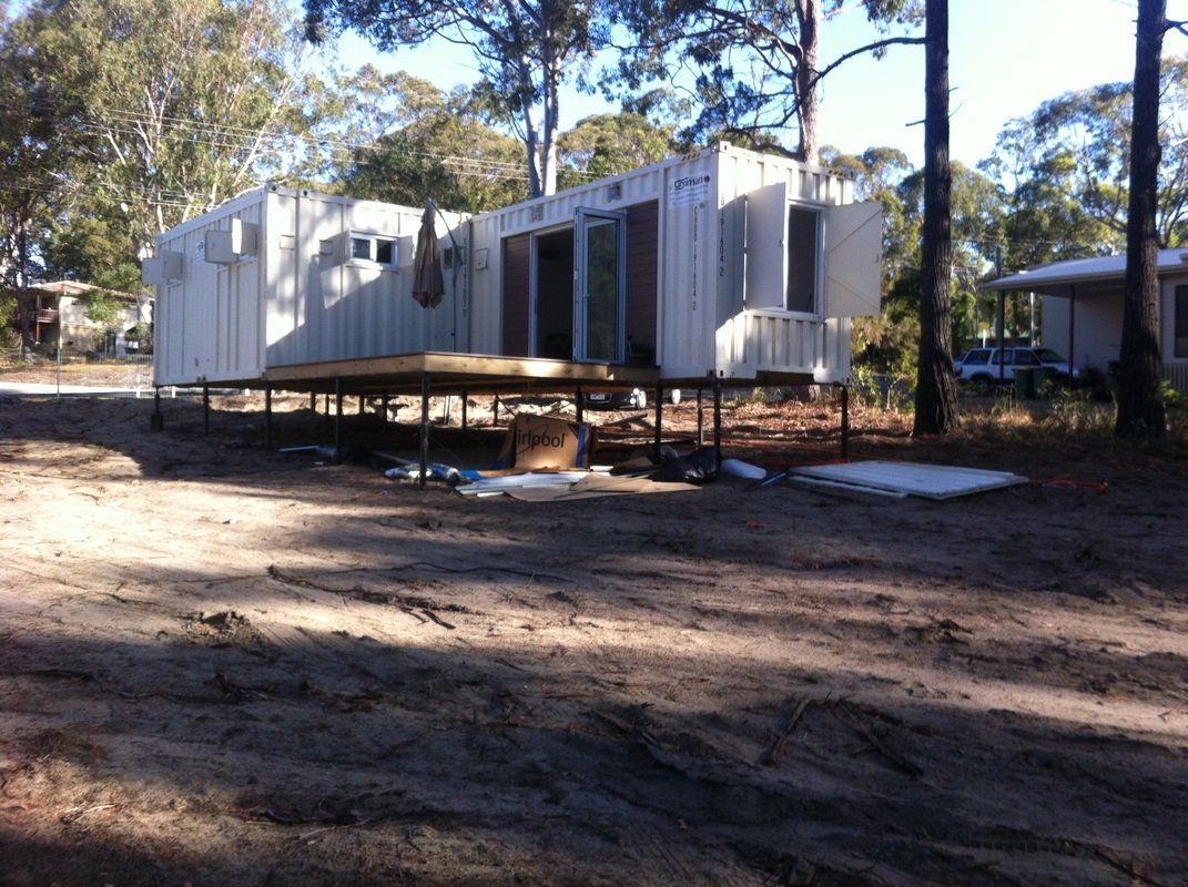 White modified shipping container raised on supports; in a dirt area, trees in the background.