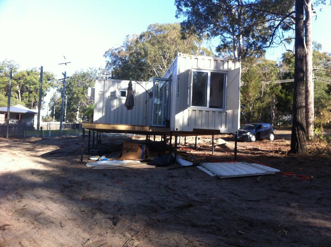 A modified shipping container house with windows and a deck outdoors on a dirt surface.