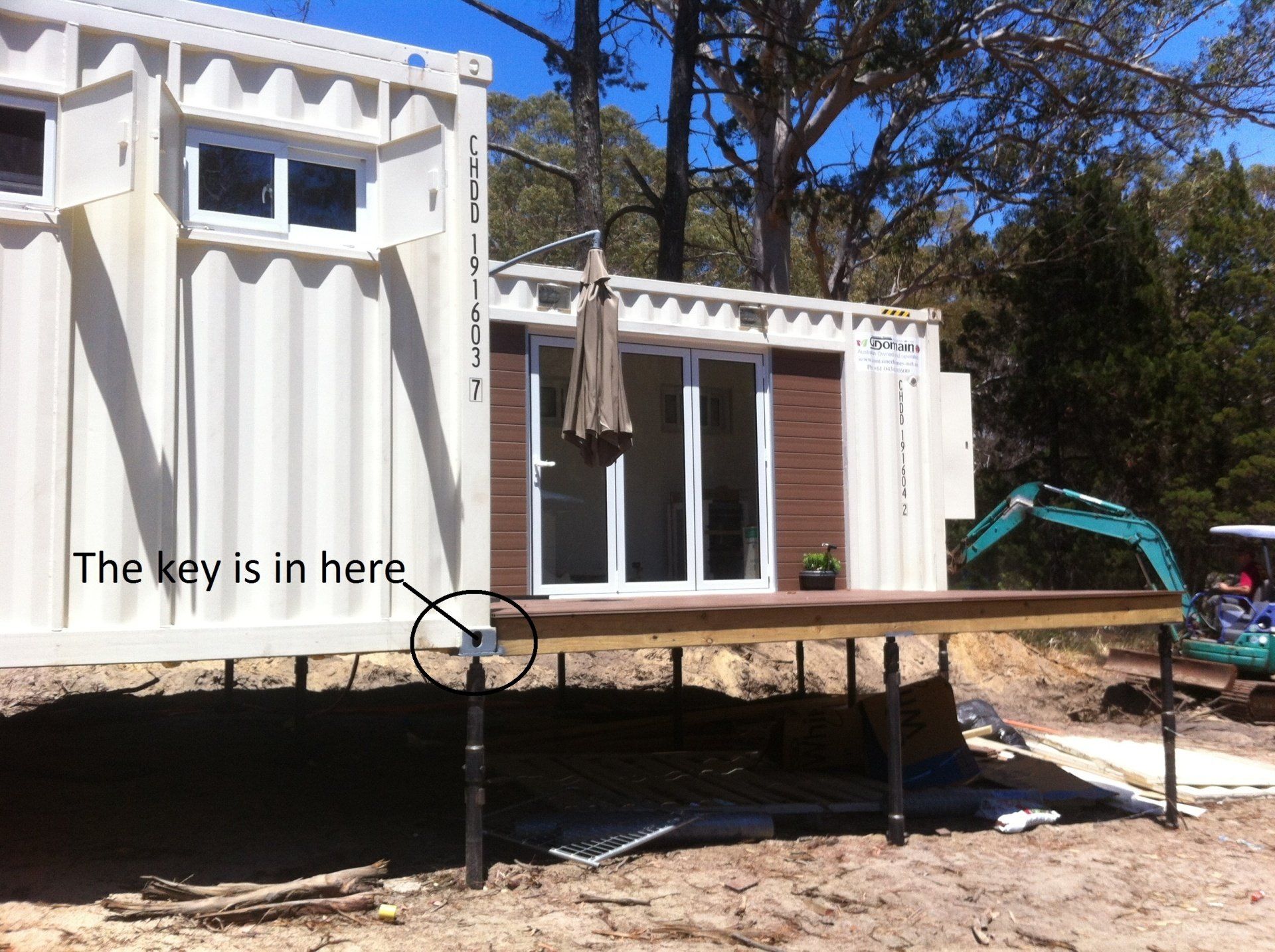 A white container home with a key visible on the deck. An excavator is also in the background.