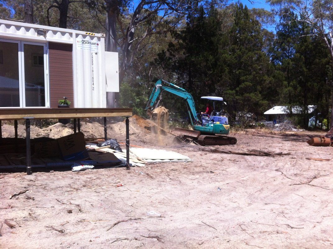 Small teal excavator moving sand near a deck and container building in a sunny, sandy area.