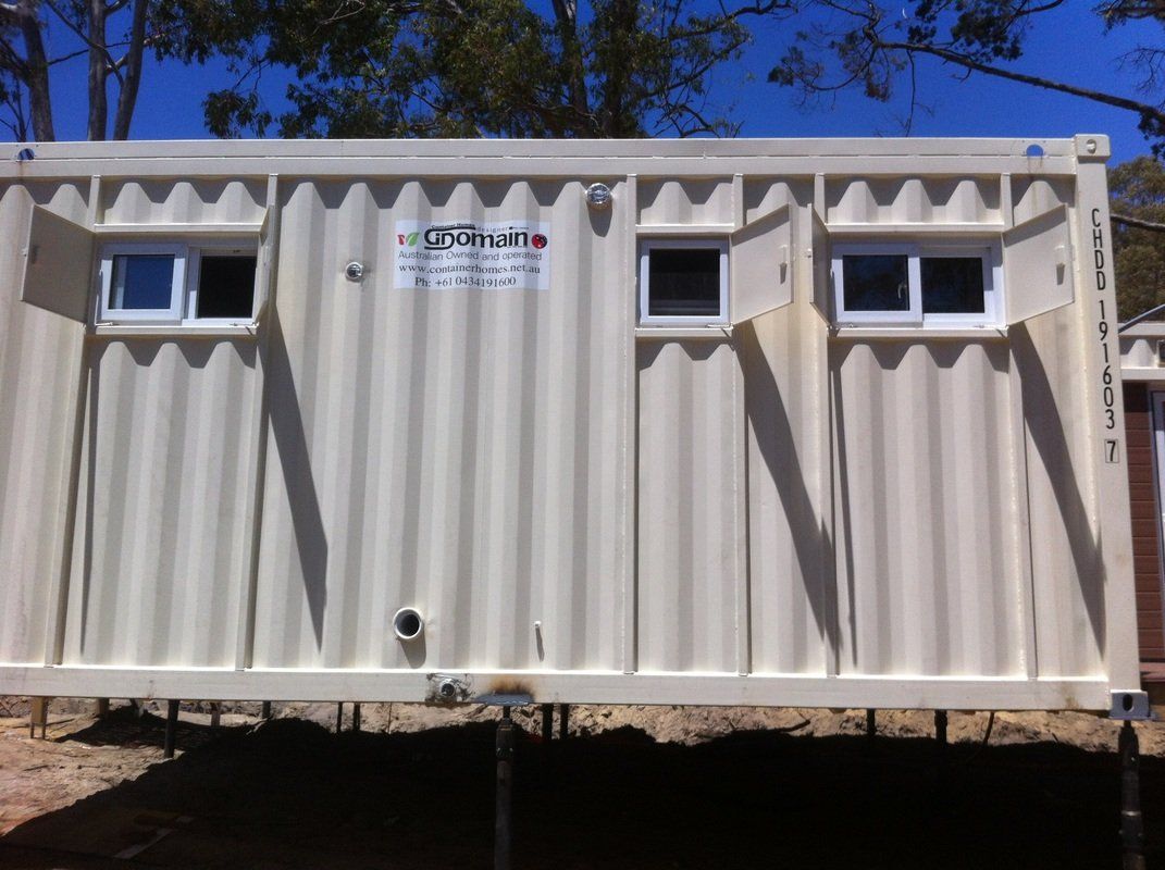Exterior view of a cream-colored shipping container modified with two windows and a small door, sitting on a raised platform.