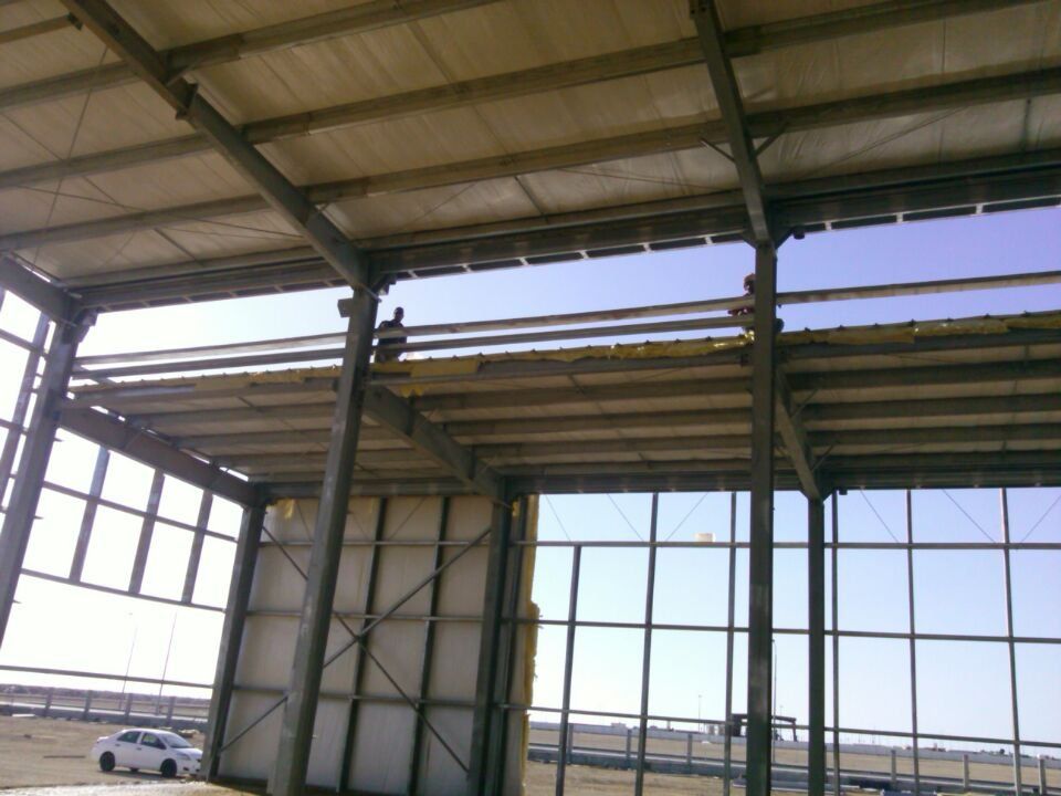 Construction workers installing panels on a metal framed building. Sunlight and blue sky are visible.