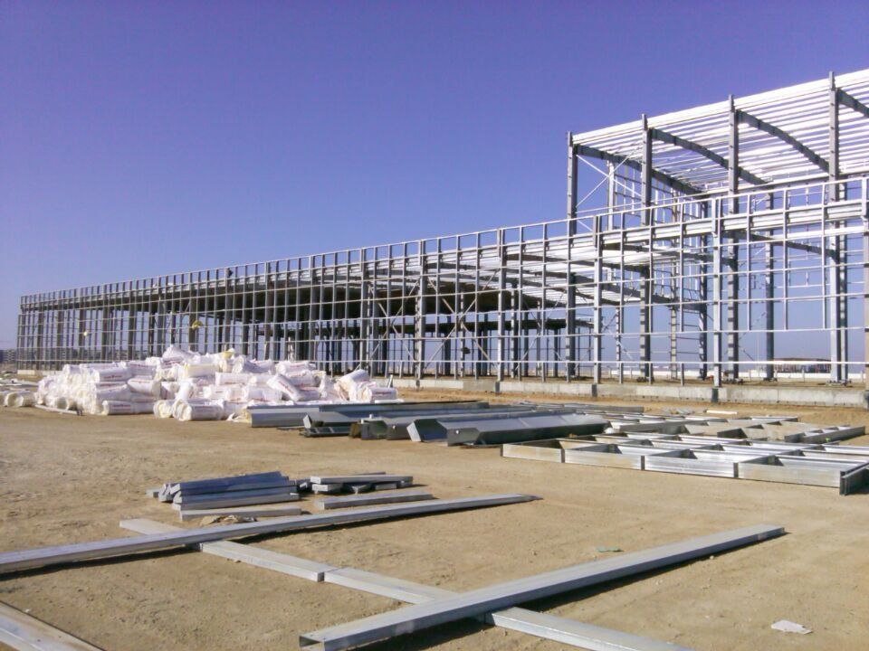 Steel-framed building under construction on a dry, open lot. Construction materials lie on the ground.