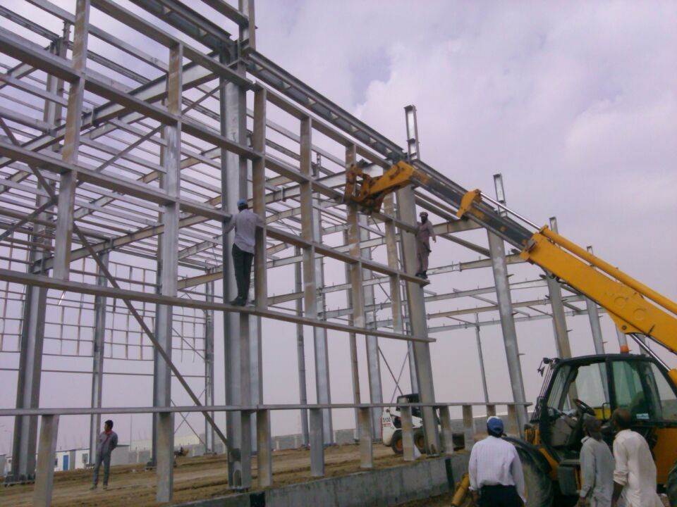 Construction workers assembling a steel frame building with a yellow forklift under a cloudy sky.