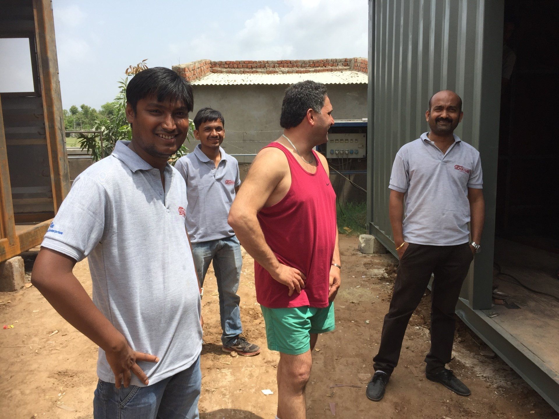 Four men standing outside a building; one in a red tank top and green shorts.