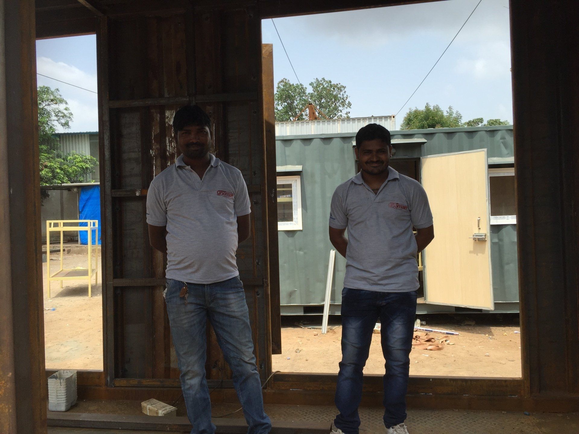 Two men in grey shirts and jeans standing in a metal frame doorway with a container building in the background.
