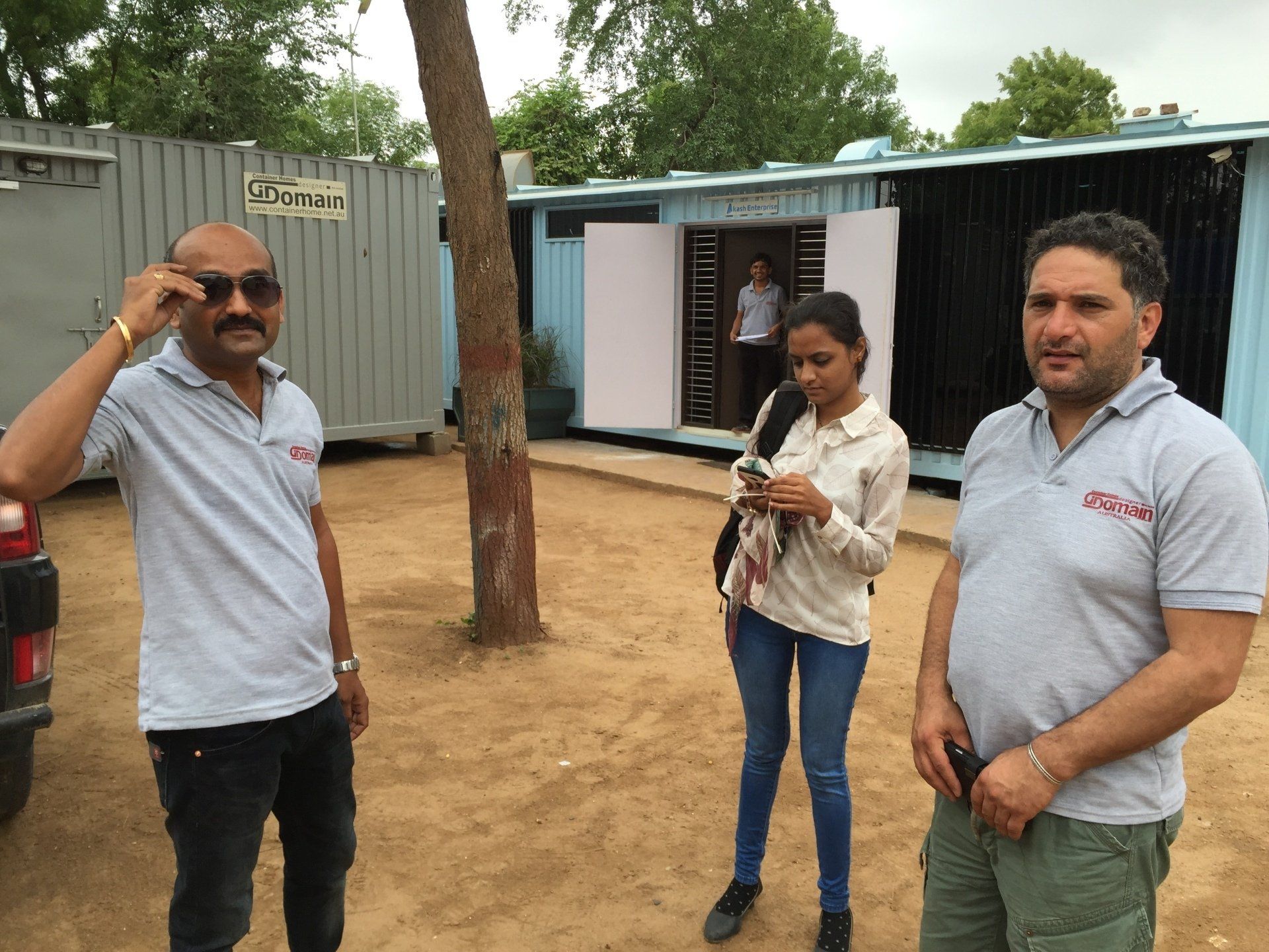 Three people standing outside shipping container buildings, one adjusting glasses, one looking at phone.