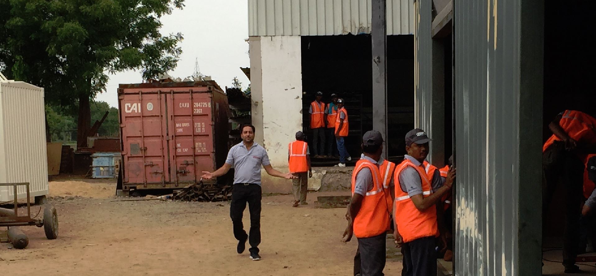 A man gestures towards a building entrance where several workers in orange vests stand, near a shipping container.