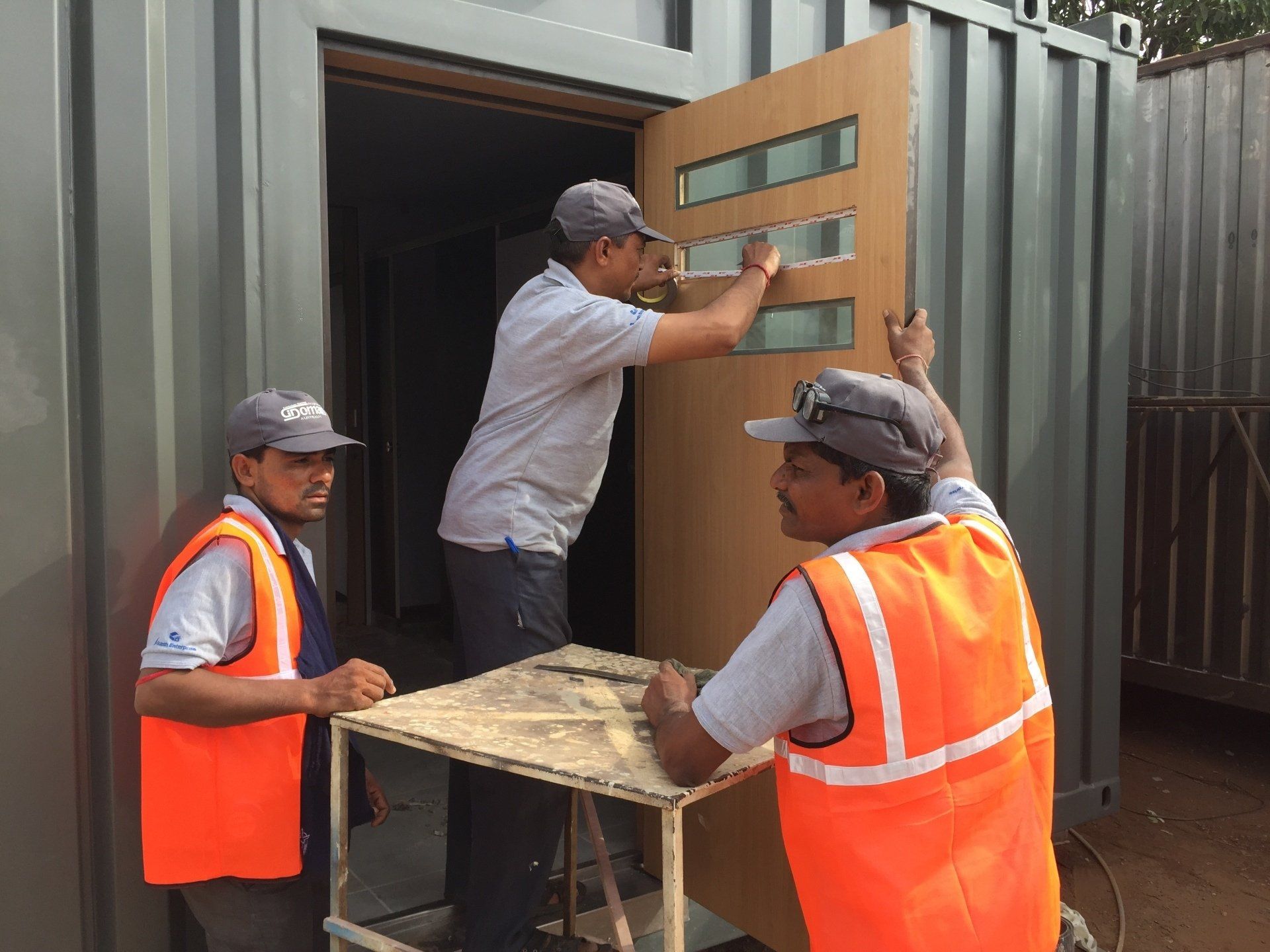 Three workers installing a door on a gray shipping container. Two workers wear orange vests and one wears a gray shirt.