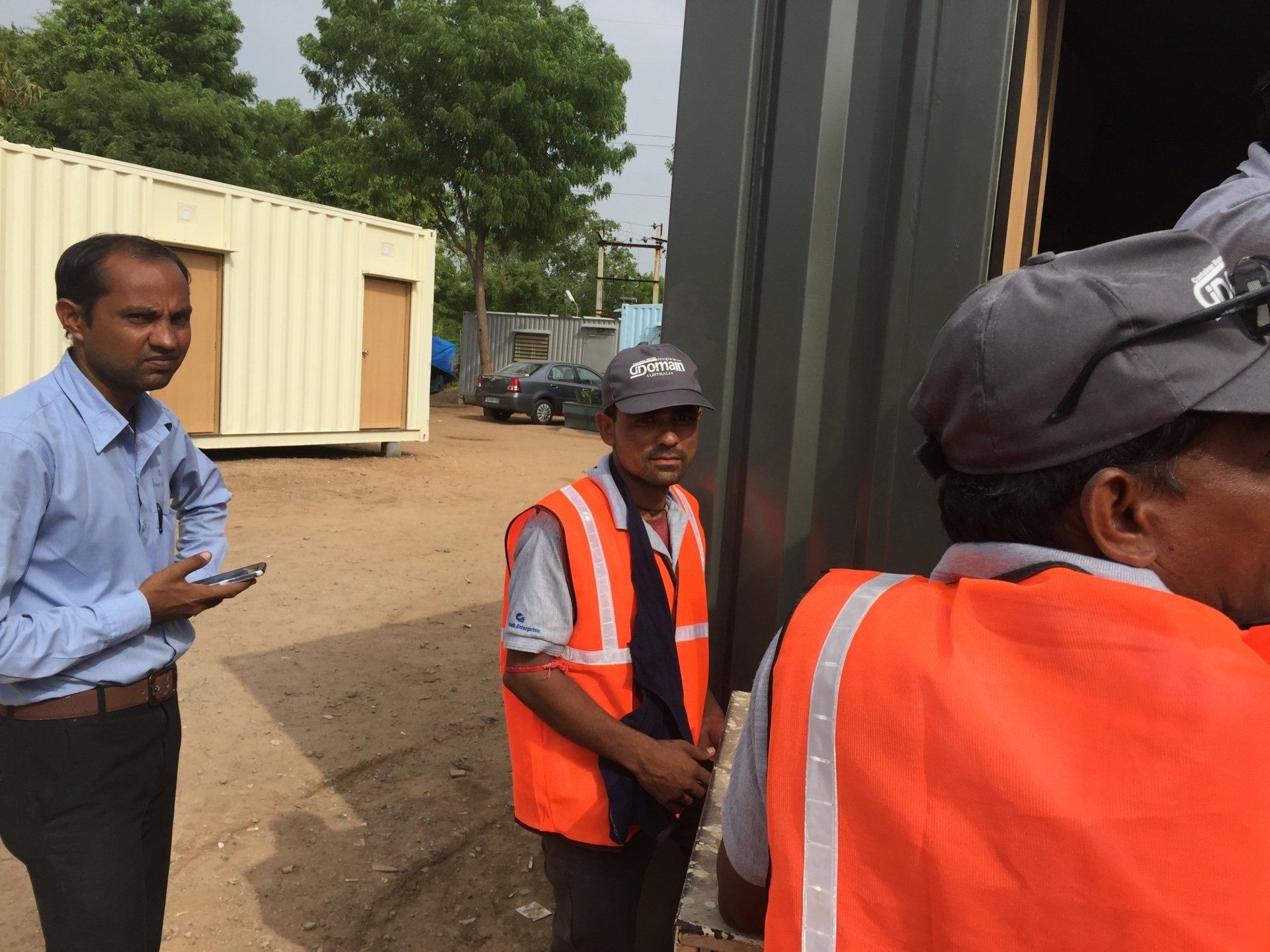 Men in work attire near shipping containers outdoors. One man holds a phone. Others wear orange vests and caps.