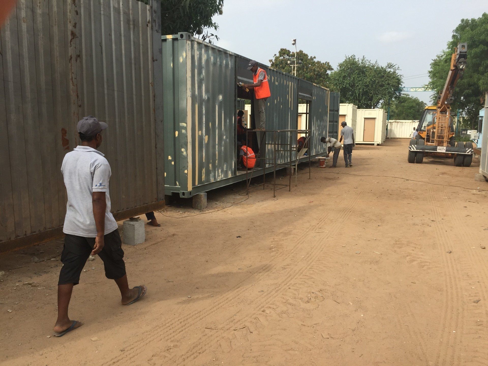 Construction workers modifying a shipping container. Outdoor setting with a forklift, other containers, and dirt ground.