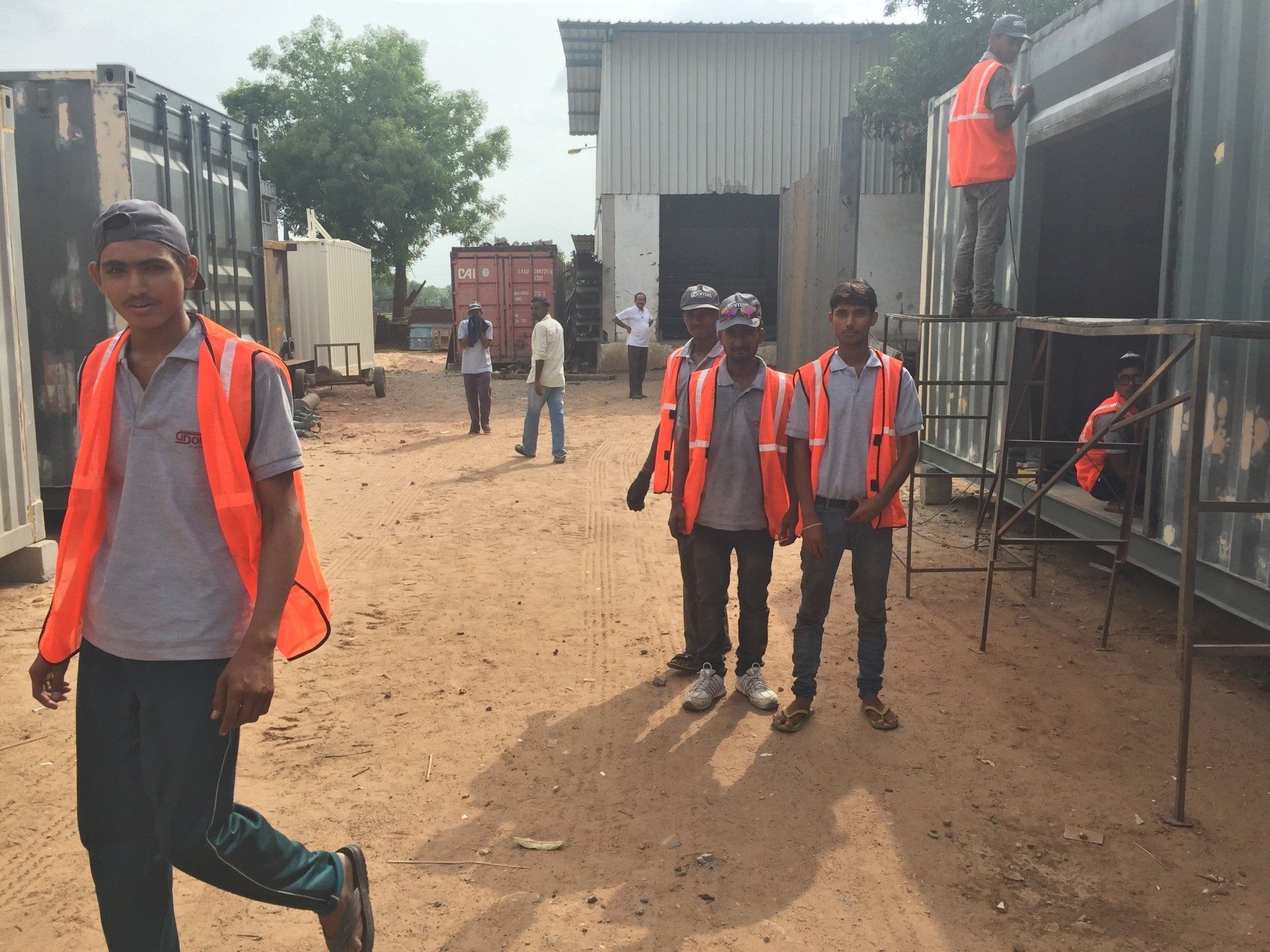 People in orange vests walking and working near shipping containers and buildings.