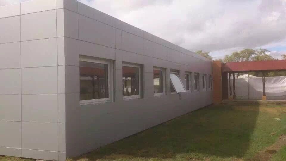 Gray building with several windows, one open, on a grassy lawn. Brown structure in the background.